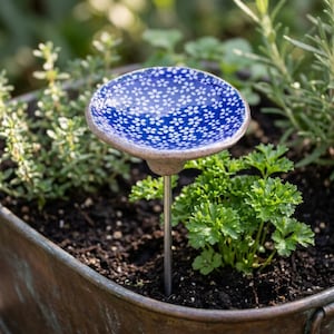 Könnte beinhalten: Ein Gartenstecker mit einer kleinen, runden und flachen Keramikschale, die eine glänzend tiefblaue Glasur und ein sich wiederholendes weißes Blumenmuster aufweist. Die Schale ist an einem dünnen, geraden, silberfarbenen Metallstab befestigt.
