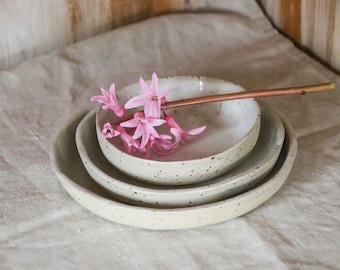 May include: A set of three speckled ceramic bowls in varying sizes, nested together. A pink flower and a copper-colored stem rest in the top bowl. The bowls are on a neutral-colored linen surface.