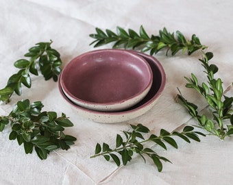 May include: Two ceramic bowls, one nested inside the other, with a speckled off-white exterior and a deep pink interior. Green leafy branches surround the bowls, arranged on a white linen surface. The bowls are ideal for serving food.