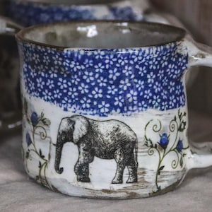 May include: Ceramic mug featuring a blue floral design and an elephant illustration. The mug has a white handle and a rustic, textured finish. Ideal for tea or coffee, this mug has a unique, handcrafted aesthetic.