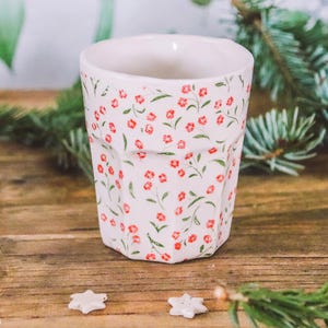 May include: A white ceramic cup with a floral pattern of small red flowers and green leaves. The cup has a faceted design and is surrounded by pine needles, pine cones, and star-shaped decorations on a wooden surface.