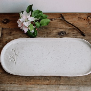 Peut inclure: Plateau de service ovale en céramique blanche avec une finition mouchetée et un motif floral. Le plateau est de forme ovale et possède un bord légèrement surélevé.