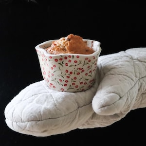Puede incluir: Un pequeño cuenco de cerámica con estampado floral lleno de productos horneados, sostenido por un guante de horno acolchado blanco. El cuenco tiene una base blanca con diseños florales rojos. El guante de horno es de color gris claro.