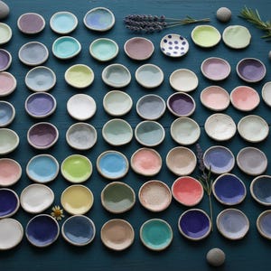 May include: A collection of small, handmade ceramic dishes in various colors, including blue, pink, green, and purple. The dishes are arranged on a dark blue surface, with sprigs of lavender and small stones scattered around them. The dishes are perfect for serving small portions.