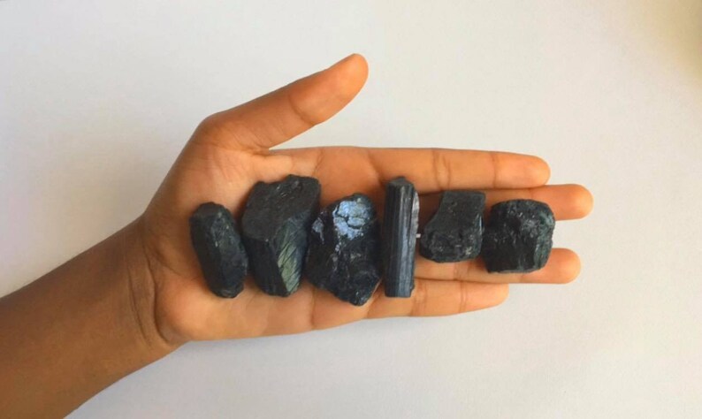 May include: A collection of black tourmaline crystals, some with a green sheen, in a hand. The crystals are various shapes and sizes.
