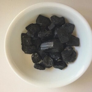 May include: A white bowl filled with black tourmaline crystals. The crystals are various shapes and sizes, with some being long and thin, while others are more rounded.