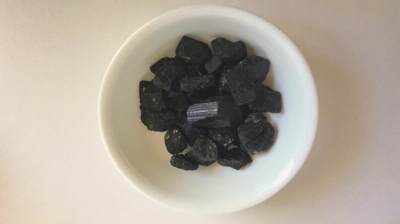 May include: A white bowl filled with black tourmaline crystals. The crystals are various shapes and sizes, with some being long and thin, while others are more rounded.