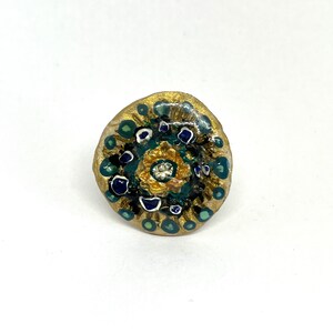 Op de afbeelding: Een ronde, gouden ring met een groen, blauw en wit geschilderd ontwerp. Het midden van de ring heeft een gouden ontwerp met een kleine, heldere steen.