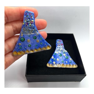 Op de afbeelding: Een paar blauwe keramische oorbellen met gouden en groene accenten. De oorbellen zijn driehoekig van vorm met een afgeronde bovenkant en een gegolfde onderkant. De oorbellen zijn versierd met een patroon van stippen en cirkels.