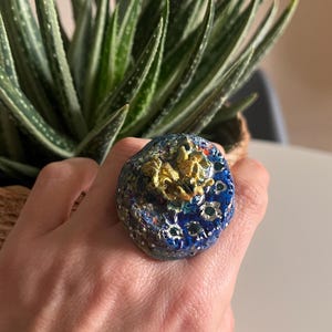 May include: A large, statement ring with a blue, gold, and white design. The ring is made of ceramic and has a textured surface.