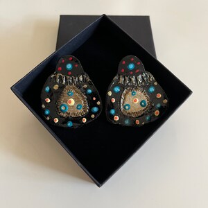 May include: A pair of black statement earrings with colourful geometric designs. The earrings are made of clay and have a textured surface. The earrings are in a black box.