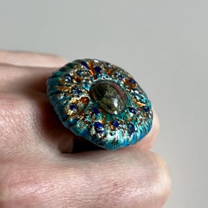 May include: A handcrafted ring with a teal, gold, and navy blue design. The ring features a central oval-shaped stone with a speckled appearance. The ring's surface has a textured, organic look, with intricate details and a unique colour palette.