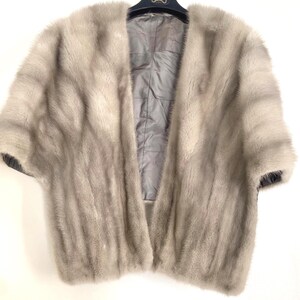 May include: A light gray fur cape with short sleeves. The cape is open in the front, revealing a gray satin lining. The garment is hanging on a black hanger. The fur appears soft and luxurious.