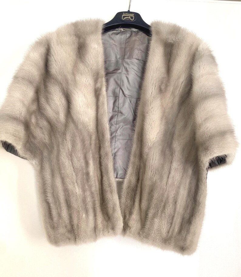 May include: A light gray fur cape with short sleeves. The cape is open in the front, revealing a gray satin lining. The garment is hanging on a black hanger. The fur appears soft and luxurious.