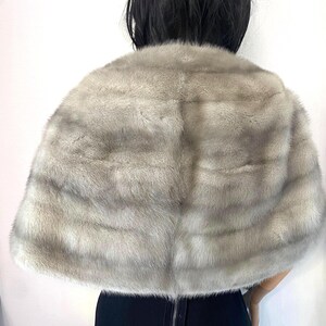 May include: A light gray fur cape with a soft, textured appearance. The cape is draped over the shoulders, with a visible back zipper on the black garment beneath. The fur has a natural, layered pattern.