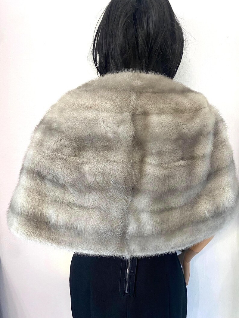 May include: A light gray fur cape with a soft, textured appearance. The cape is draped over the shoulders, with a visible back zipper on the black garment beneath. The fur has a natural, layered pattern.