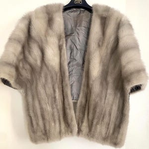 May include: A light gray fur cape with short sleeves, hanging on a black hanger. The interior lining is a quilted gray fabric. The fur appears soft and plush, with a natural, flowing texture.