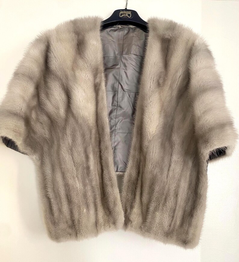 May include: A light gray fur cape with short sleeves, hanging on a black hanger. The interior lining is a quilted gray fabric. The fur appears soft and plush, with a natural, flowing texture.