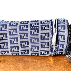 May include: A cylindrical handbag with a blue and grey repeating pattern. The bag has a silver zip and a matching handle. The bag is on a wooden surface.