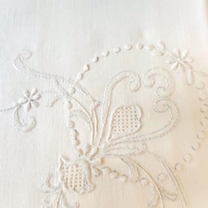 May include: White linen fabric with delicate white embroidery of a floral design. The embroidery features intricate swirls and a small flower.
