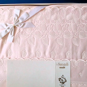 Puede incluir: Una colcha bordada de color rosa con un lazo de cinta de satén blanco y una etiqueta blanca con el texto "i Sangalli" en letras negras.