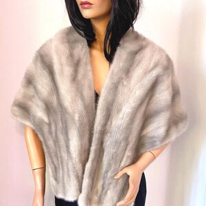 May include: A light gray fur shawl draped over a mannequin. The shawl has a soft, textured appearance and a wide, open design. The mannequin is wearing a black top and a dark blue skirt.