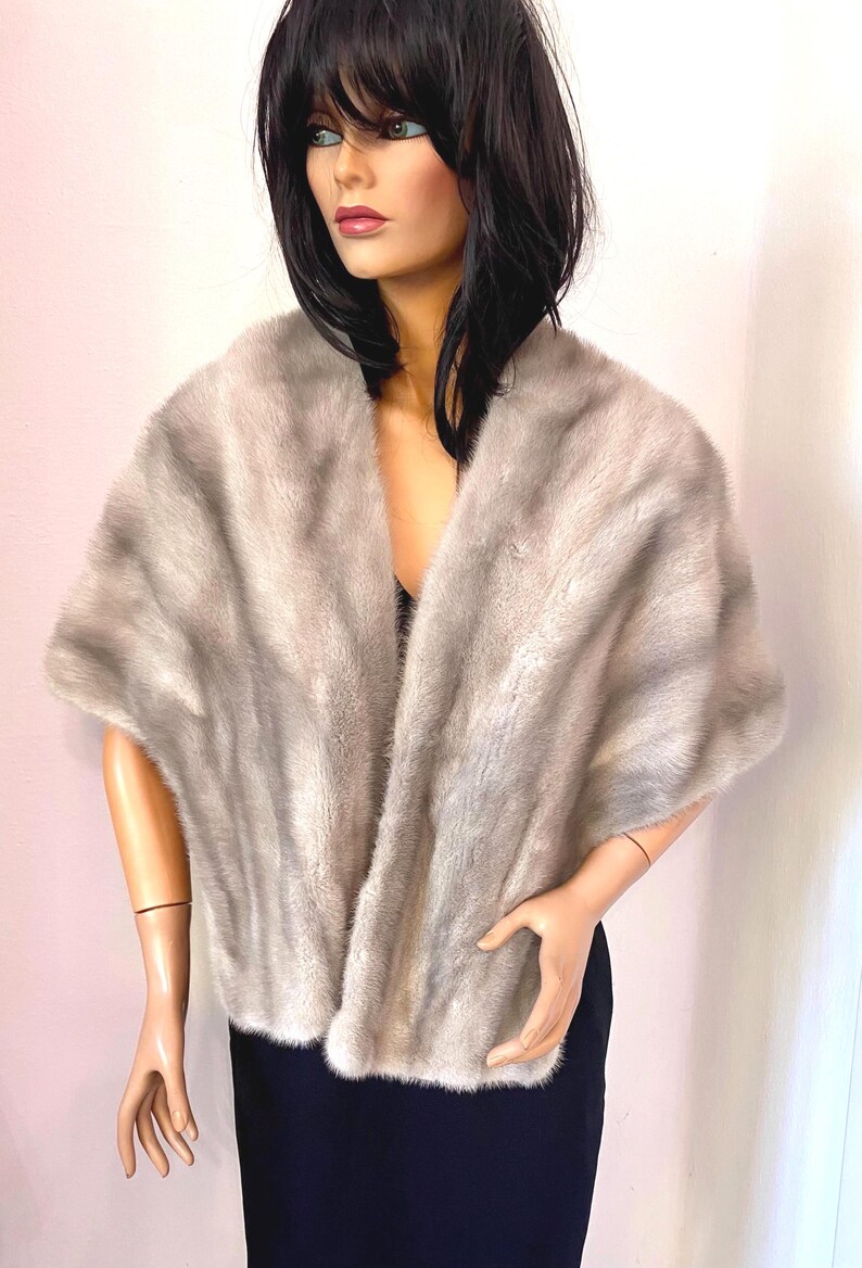May include: A light gray fur shawl draped over a mannequin. The shawl has a soft, textured appearance and a wide, open design. The mannequin is wearing a black top and a dark blue skirt.