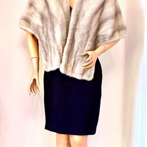 May include: A light gray fur stole draped over a black dress on a mannequin. The fur has a soft, textured appearance. The mannequin has dark hair and is posed in front of a white wall. The dress is knee-length.