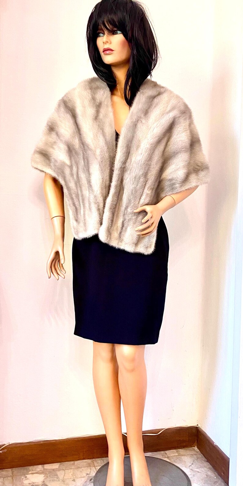 May include: A light gray fur stole draped over a black dress on a mannequin. The fur has a soft, textured appearance. The mannequin has dark hair and is posed in front of a white wall. The dress is knee-length.