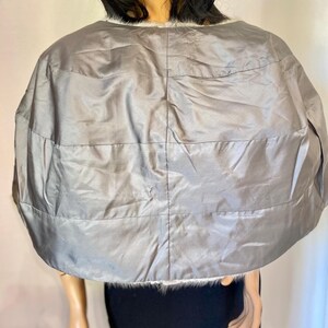 May include: A silver-gray cape with horizontal stitched lines. The cape has a rounded shape and is trimmed with white faux fur along the neckline. The garment appears to be made of a shiny, quilted material.
