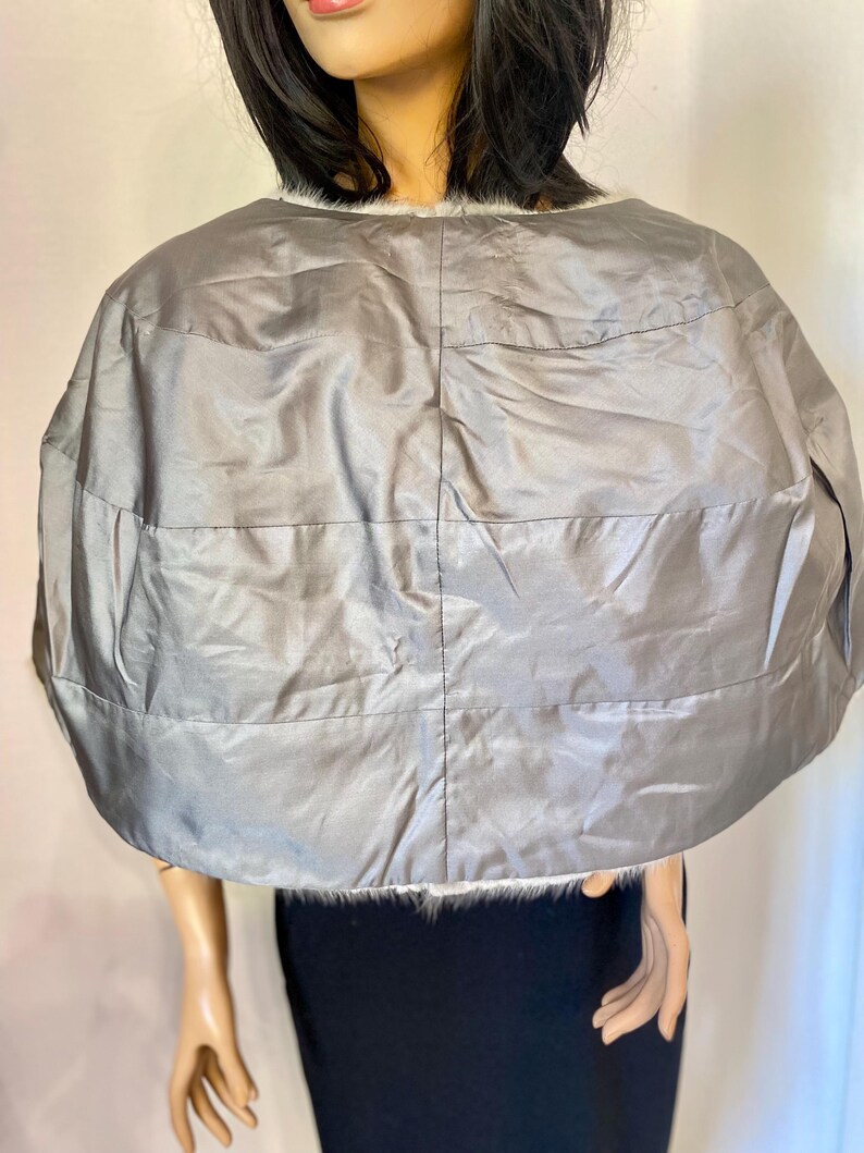 May include: A silver-gray cape with horizontal stitched lines. The cape has a rounded shape and is trimmed with white faux fur along the neckline. The garment appears to be made of a shiny, quilted material.