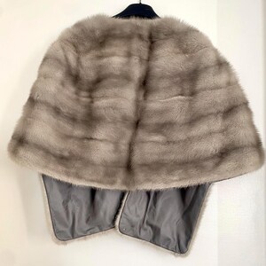 May include: A light gray fur cape with a dark gray lining. The cape is hanging on a black hanger against a white background. The fur has horizontal stripes and a soft texture.