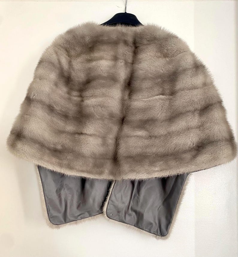 May include: A light gray fur cape with a dark gray lining. The cape is hanging on a black hanger against a white background. The fur has horizontal stripes and a soft texture.