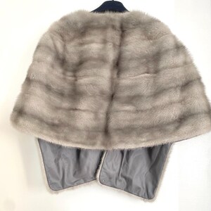 May include: A light gray fur cape with a dark blue collar and a gray satin lining. The cape is hanging on a hanger against a white wall. The fur has horizontal stripes.