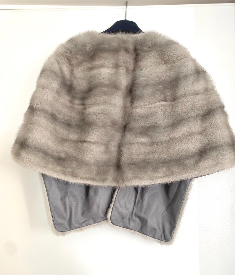 May include: A light gray fur cape with a dark blue collar and a gray satin lining. The cape is hanging on a hanger against a white wall. The fur has horizontal stripes.