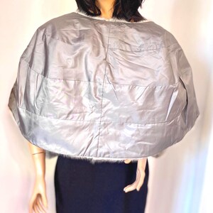 May include: A silver quilted cape with a faux fur trim. The cape is a light gray color and has horizontal stitching. The cape is worn over a black dress. Fashion accessory.