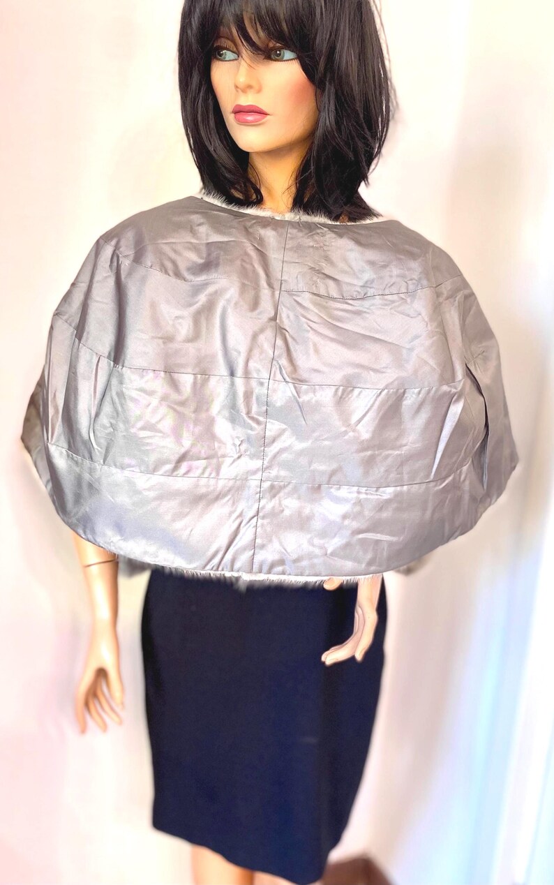 May include: A silver quilted cape with a faux fur trim. The cape is a light gray color and has horizontal stitching. The cape is worn over a black dress. Fashion accessory.