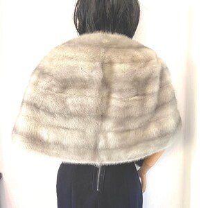 May include: A light gray fur cape with horizontal stripes. The cape is draped over a person wearing a black outfit. The fur appears soft and luxurious, with a subtle sheen. The cape is a classic fashion accessory.