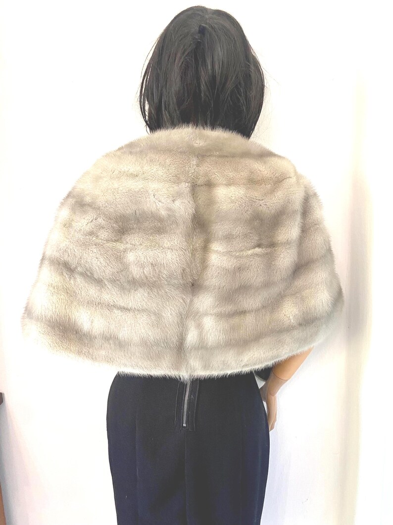 May include: A light gray fur cape with horizontal stripes. The cape is draped over a person wearing a black outfit. The fur appears soft and luxurious, with a subtle sheen. The cape is a classic fashion accessory.