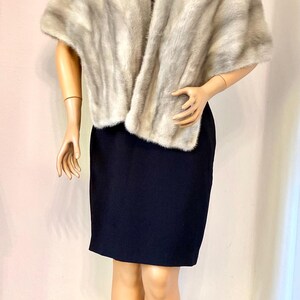 May include: A light gray fur shawl draped over a black dress on a mannequin. The shawl has a soft, textured appearance. The mannequin is standing on a gray circular base. The background is a plain wall.