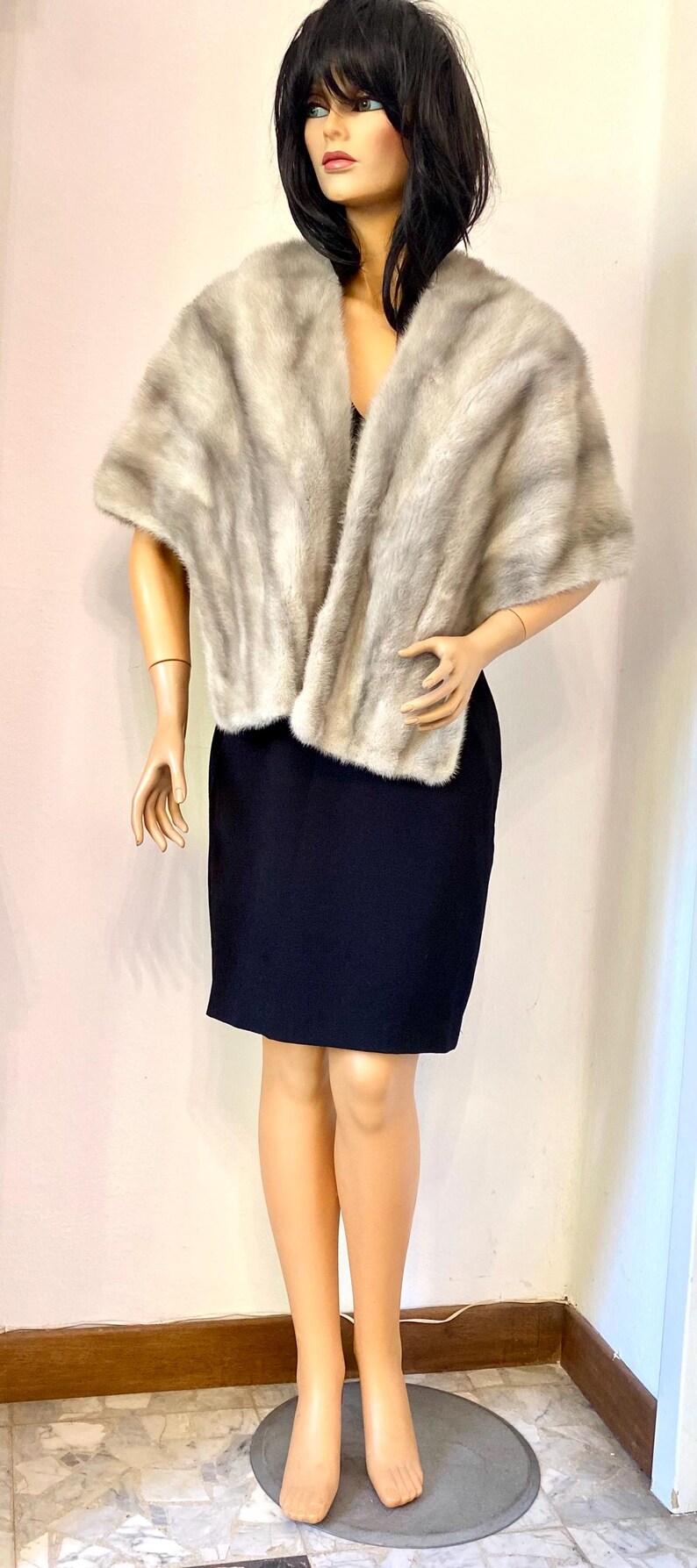 May include: A light gray fur shawl draped over a black dress on a mannequin. The shawl has a soft, textured appearance. The mannequin is standing on a gray circular base. The background is a plain wall.