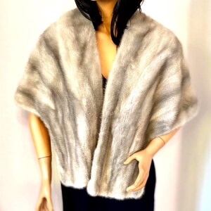 May include: A light gray faux fur shawl with a soft, textured appearance. The shawl is draped over a mannequin wearing a black dress. The fur has a gradient effect, transitioning from lighter to darker shades of gray.