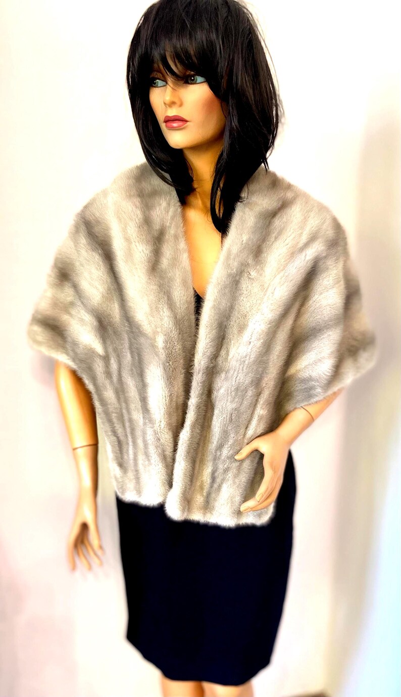 May include: A light gray faux fur shawl with a soft, textured appearance. The shawl is draped over a mannequin wearing a black dress. The fur has a gradient effect, transitioning from lighter to darker shades of gray.