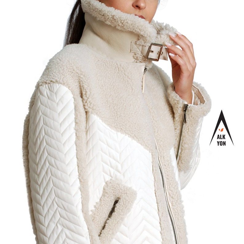 May include: A white faux fur jacket with a quilted white fabric panel on the sleeves and a zipper closure. The jacket has a high collar and a buckle detail on the collar.