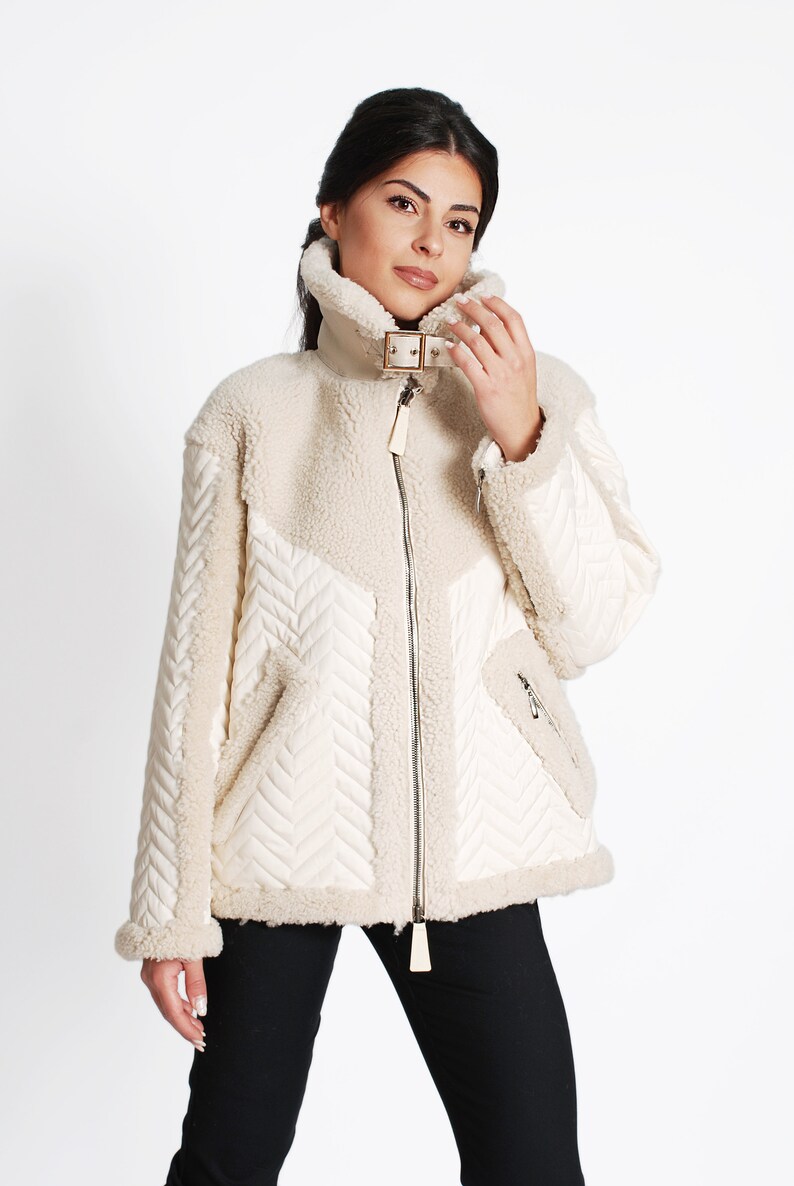 May include: A cream-colored, faux fur jacket with a quilted design. The jacket has a zipper closure and a buckle detail on the collar.
