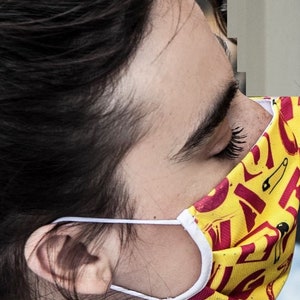 May include: A person wearing a yellow and red face mask with a white elastic band. The mask has a pattern of safety pins and the word "SAFE" repeated on it.
