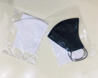 Puede incluir: Dos mascarillas, una blanca y una negra, están empaquetadas en bolsas de plástico transparentes. Las mascarillas están hechas de tela y tienen lazos elásticos para las orejas.