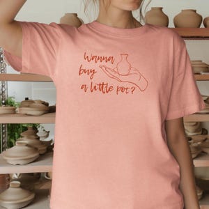 May include: A pink t-shirt with the text "Wanna buy a little pot?" in red. A hand is holding a small pot in the text.