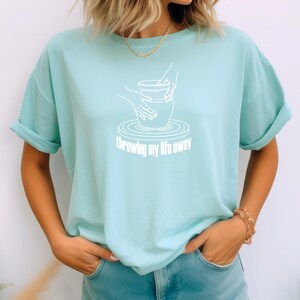 Puede incluir: Una camiseta azul claro con un gráfico blanco de una mano lanzando arcilla en un torno de alfarería. El texto "throwing my life away" está impreso debajo del gráfico.