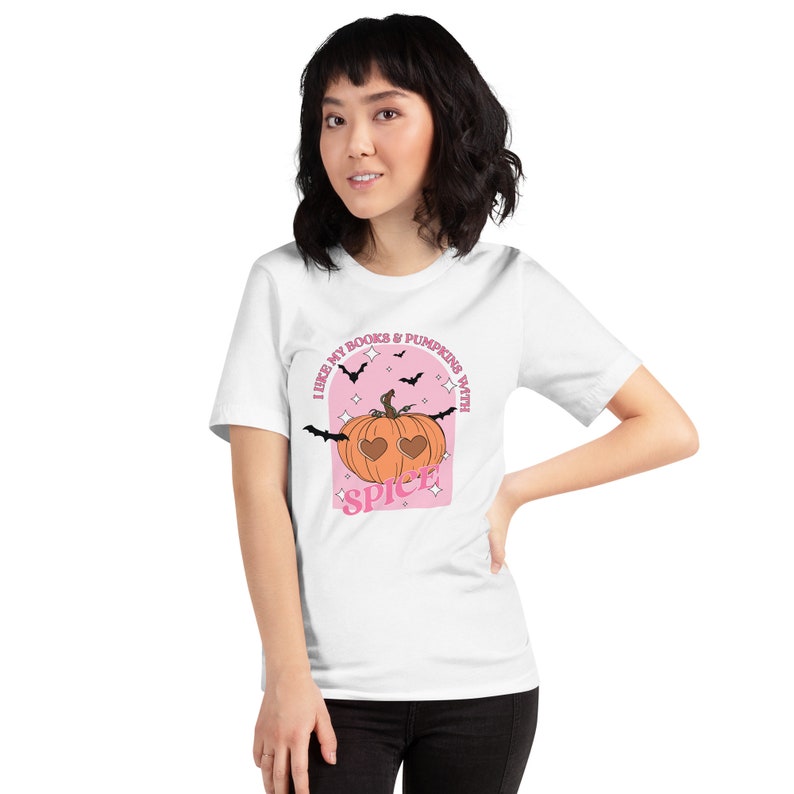 May include: White t-shirt with a pink and orange pumpkin graphic. The pumpkin has heart-shaped eyes and the text "I like my books & pumpkins with spice" is written around it.